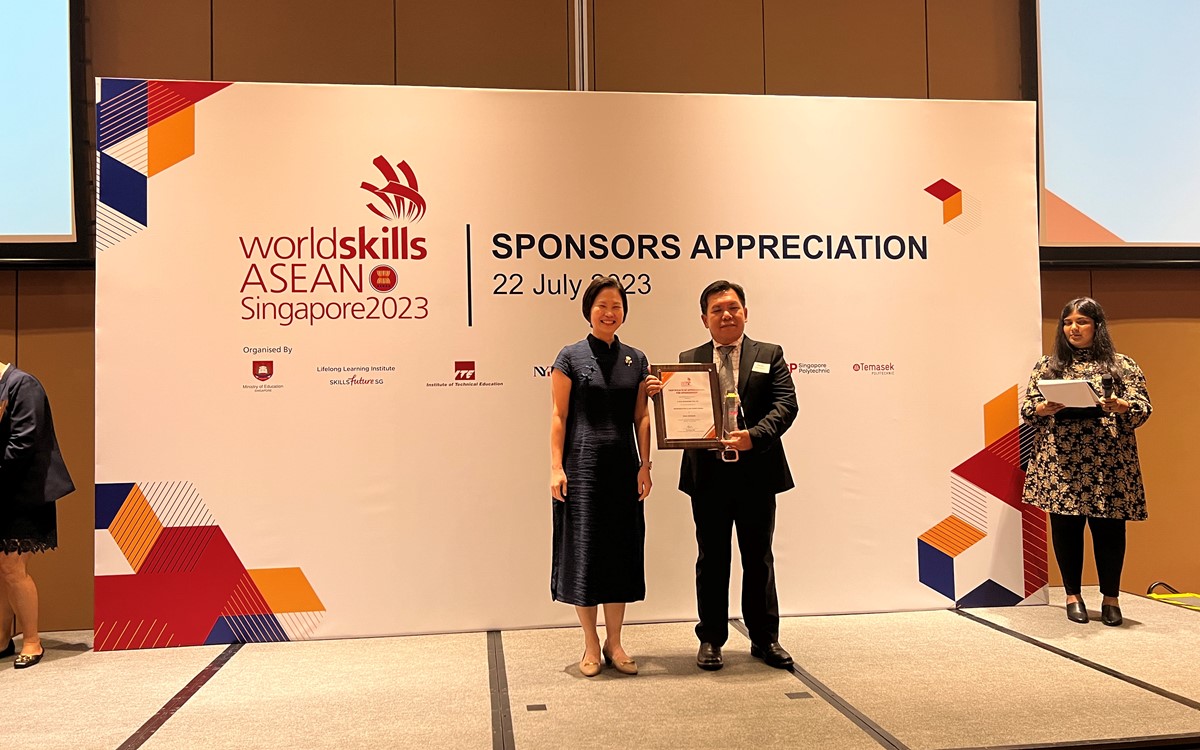Two smartly dressed people hold an award while standing in front of a white banner onstage