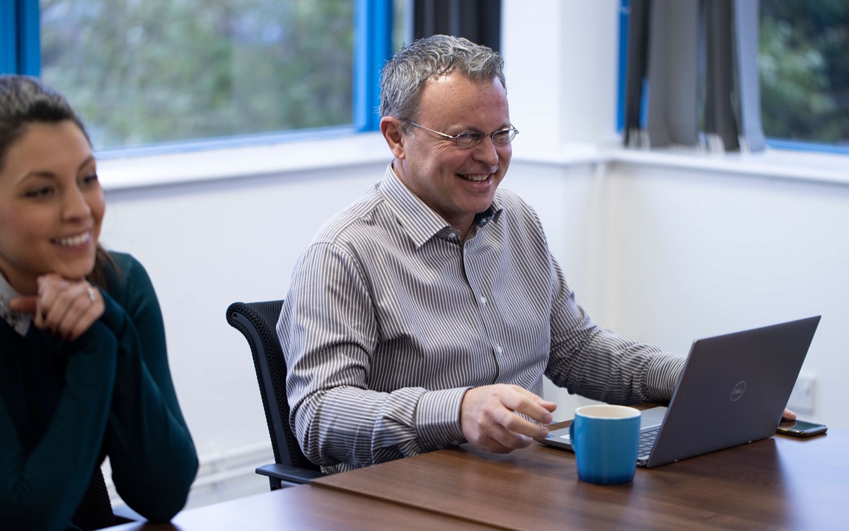 A man smiling sat a laptop with a mug of a hot drink with him