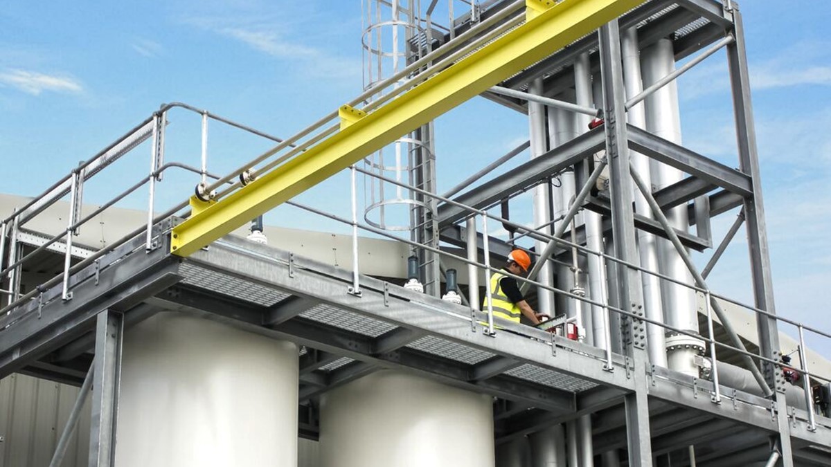 A technician in a yellow high-vis jacket and hardhat works on scaffolding on top of two white separation towers