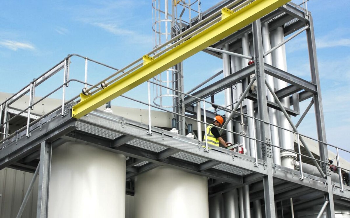 A technician in a yellow high-vis jacket and hardhat works on scaffolding on top of two white separation towers