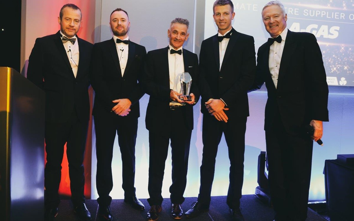 A group of smartly dressed men stand onstage and hold an award while looking at the camera