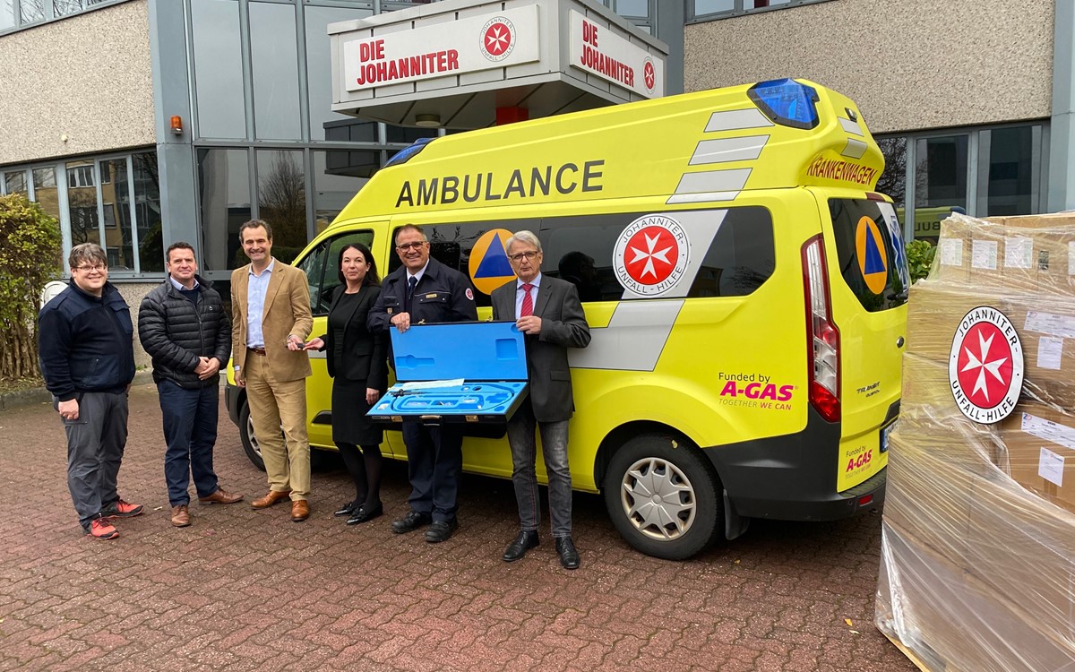 A group of people stand in front of yellow ambulance holding an open blue box