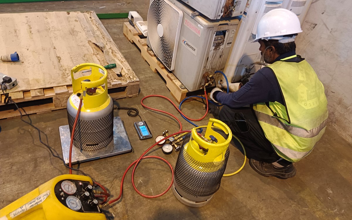 A man in a yellow high-vis jacket recovers refrigerant from grey air conditioning units in a warehouse