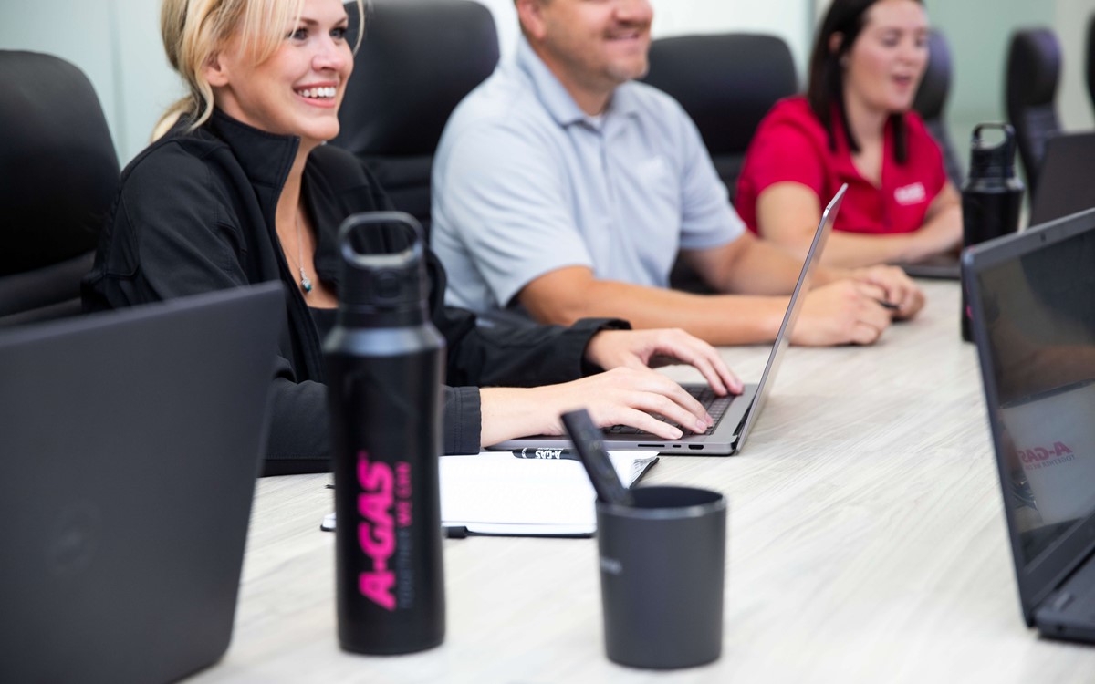 Three people sat at a boardroom desk with laptops, they are all smiling