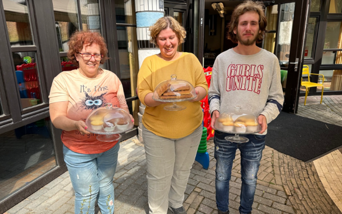Two women and a man hold trays of cakes in a courtyard in front of a building