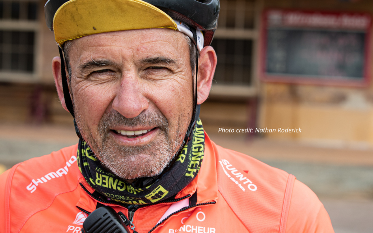 A man in an orange cycling jersey and a cycling helmet smiles at the camera outside