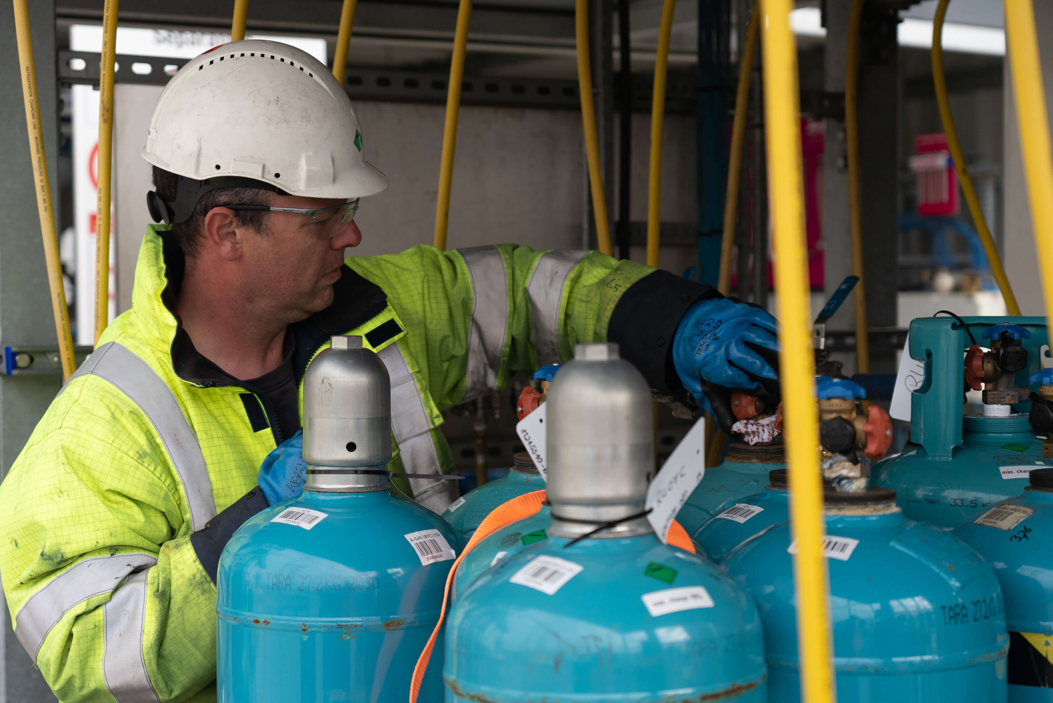 A man in a helmet and yellow hi-vis jacket works on blue cylinders 