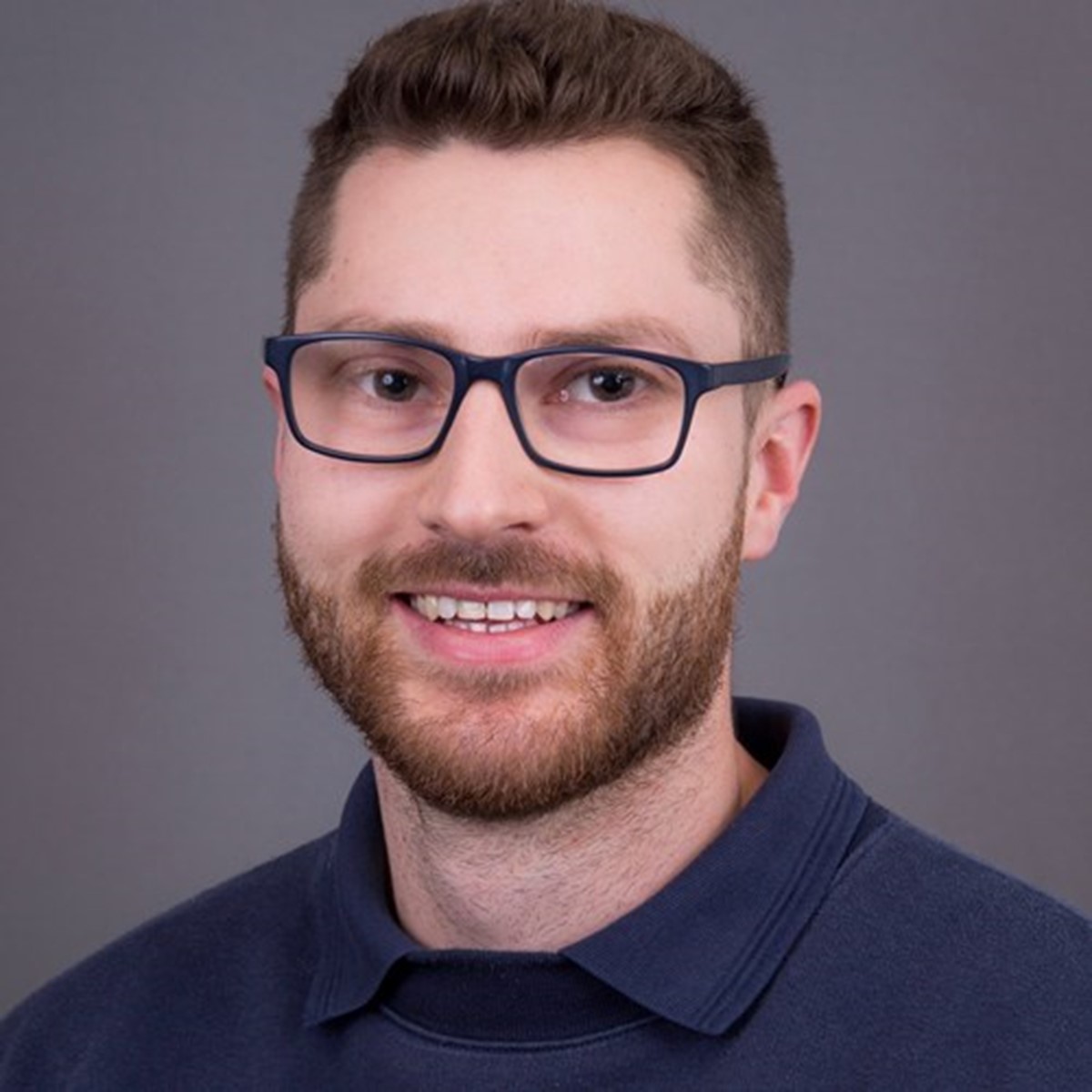 A headshot of a man in a blue polo and glasses smiling on a grey background