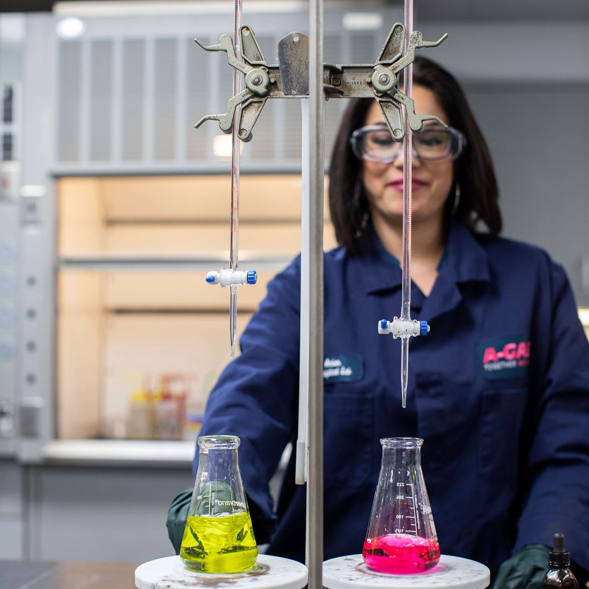 A woman carrying out laboratory analysis, she is wearing a navy lab coat and safety glasses, she is working with two liquids in scientific beakers, one is yellow and one is pink