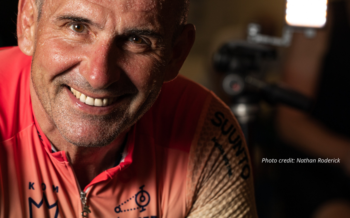 A man in an orange cycling jersey smiles at the camera against a dark background
