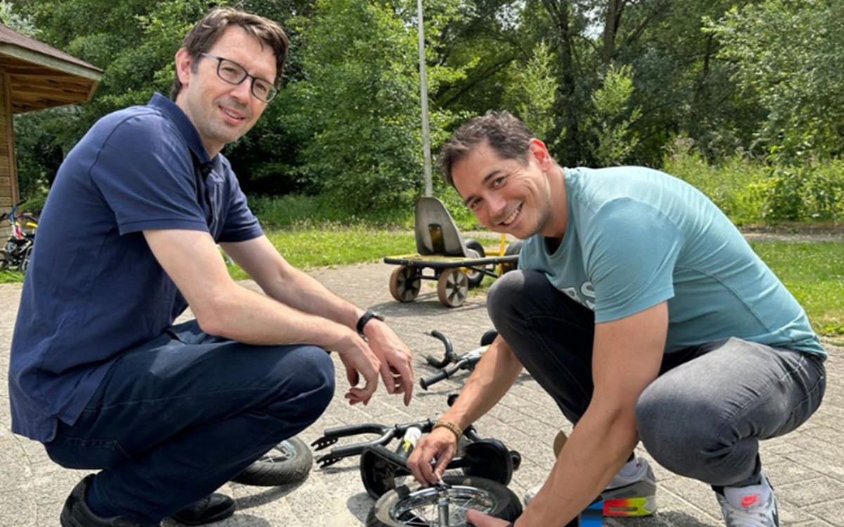 Two casually dressed men smile at the camera while fixing a bicycle outside