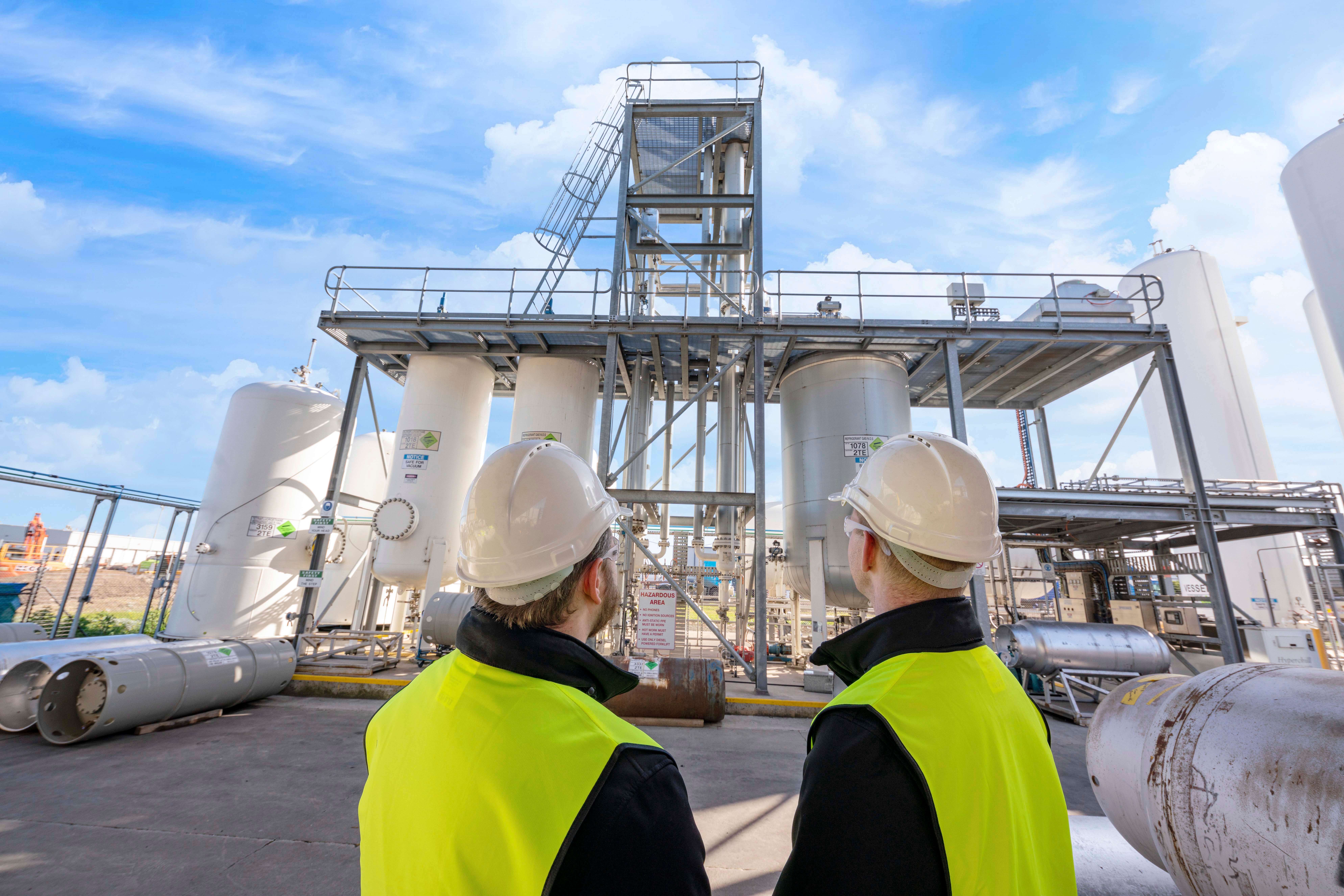 Two men in yellow high-vis jackets looking at white separation towers and metal scaffolding 