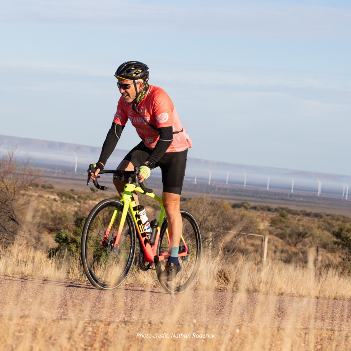 A man in an orange jersey cycling on a road across some grassland