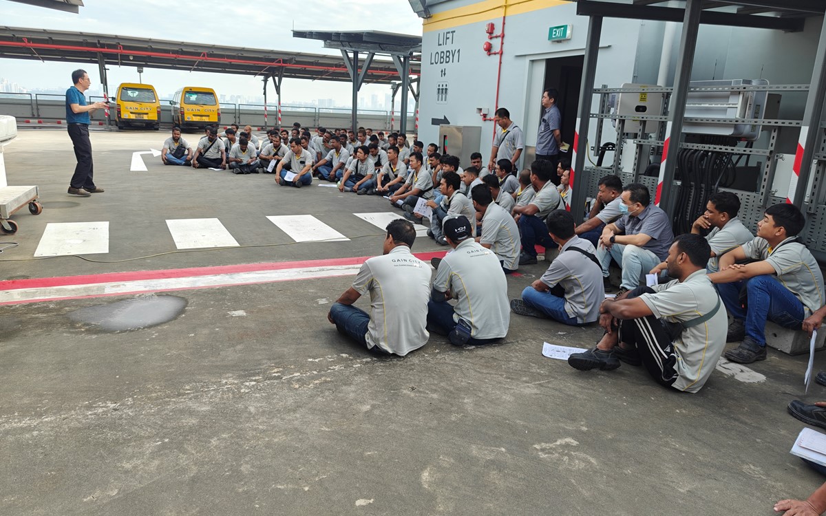 A wide shot of a man speaking to a group of people sitting down on-side