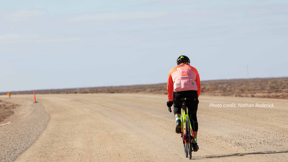 A person in an orange jersey cycling along a desert road from behind