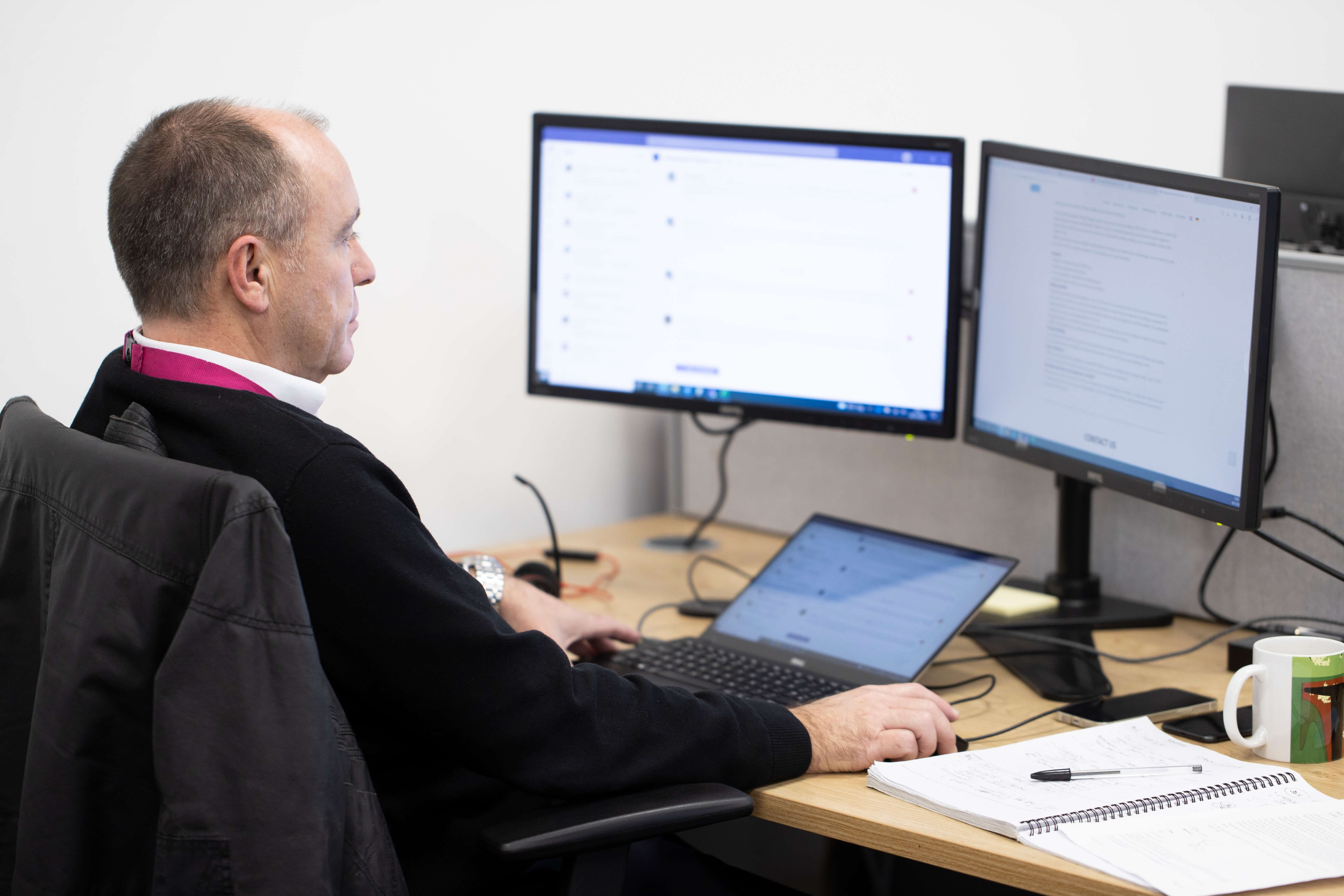 A man sat at his computer screens. The image is taken from behind so we can see him looking at his screens