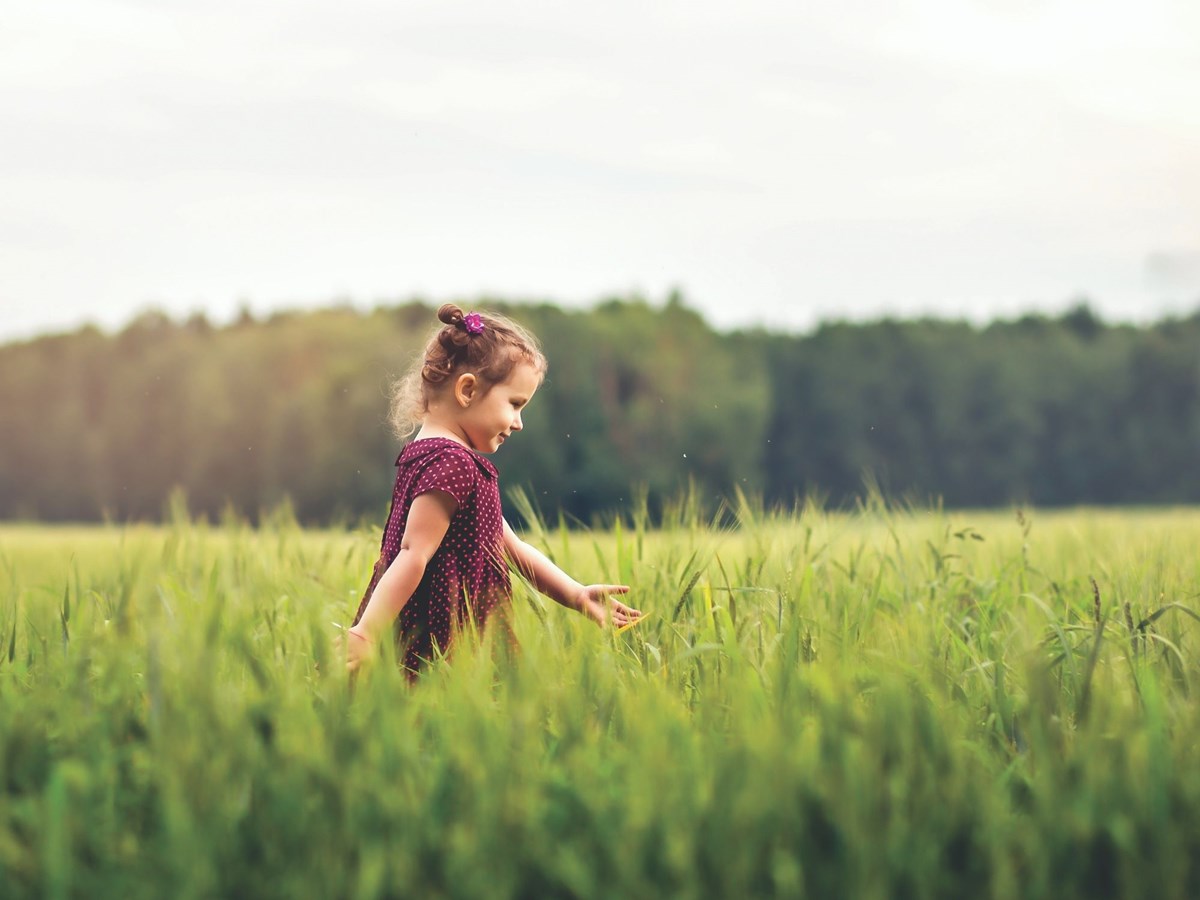 young girl walking left to right through a green field with forest in background
