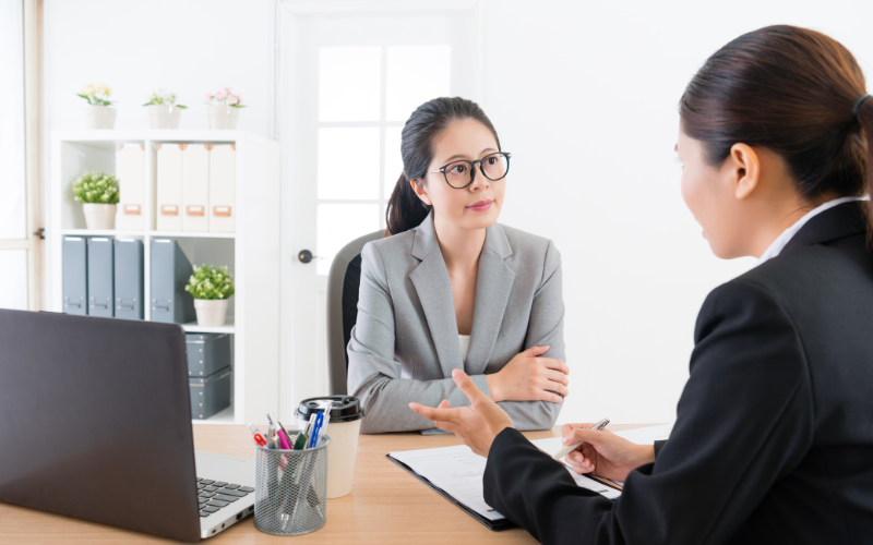 Two formally dressed women speaking at a desk in an office 