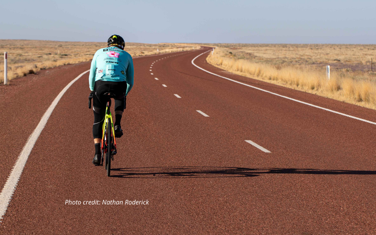 A person in a light blue jersey cycling along a desert road from behind