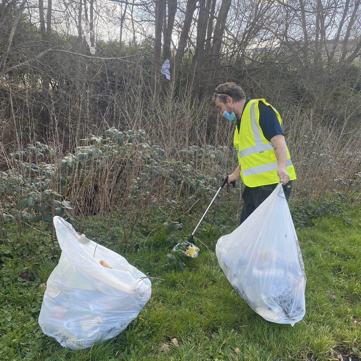 A man in a yellow high-vis jacket picks up rubbish with a litter picker in a wooded area