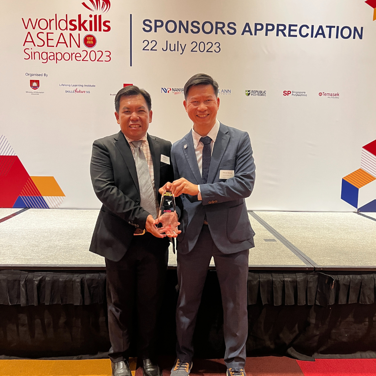 Two men smile while holding an award and looking at the camera in front of a white banner