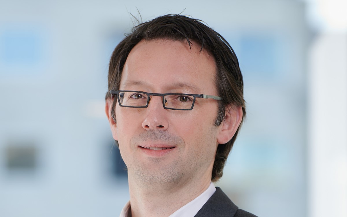 A headshot of a smartly dressed man wearing glasses smiling against a blurred background