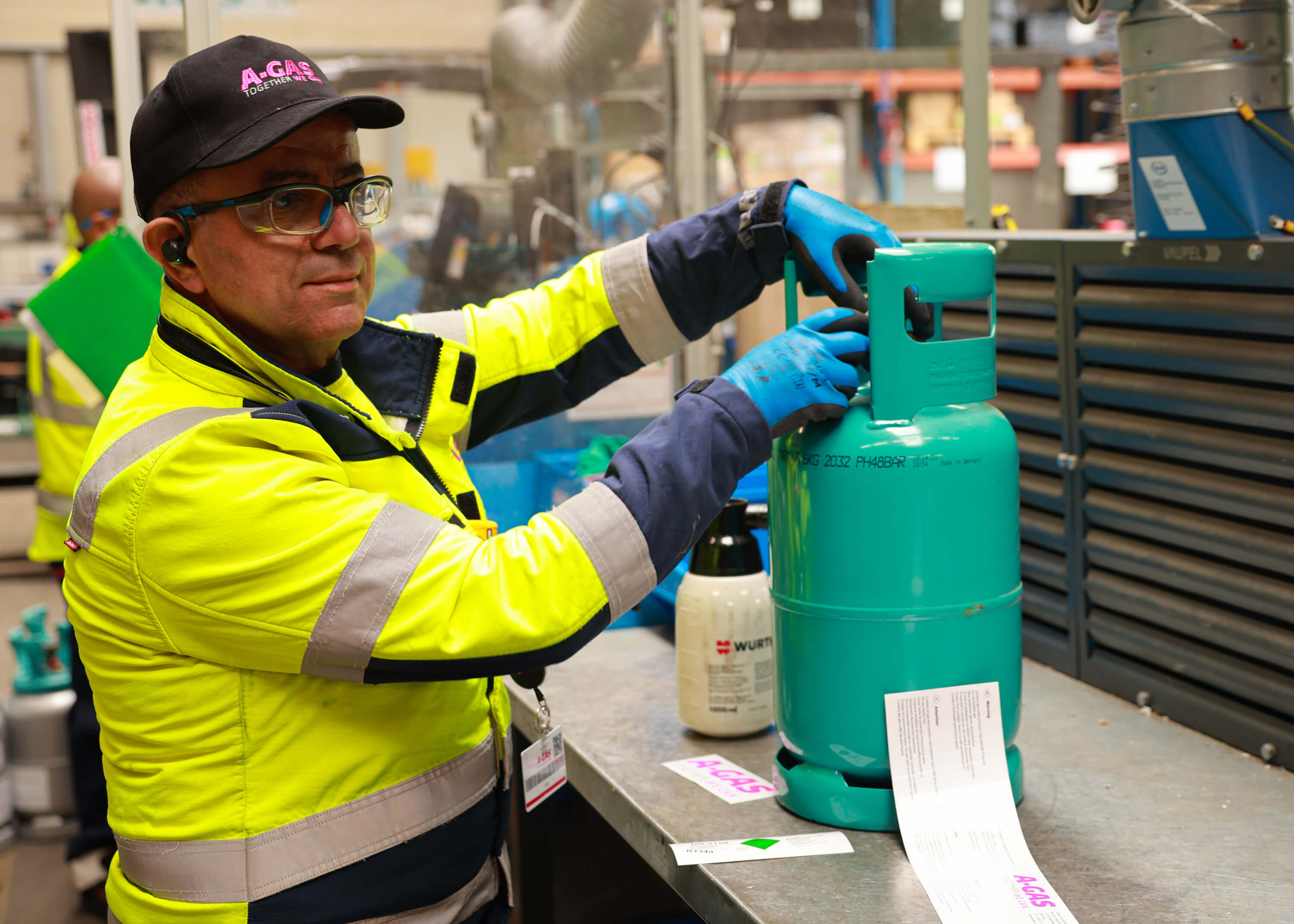 A man in a yellow high-vis jacket works with a valve on a blue cylinder in a warehouse 