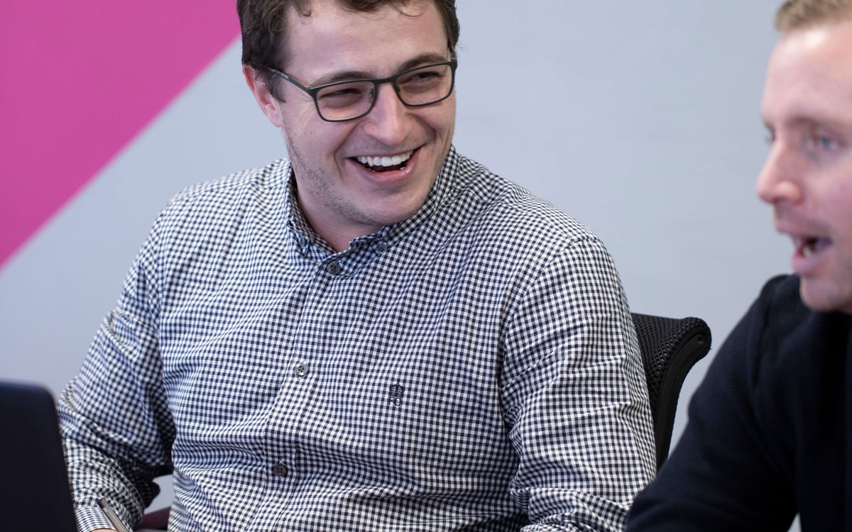 A man sat at a desk laughing, he has a laptop in front of him and is looking at his male colleague who is half in the image