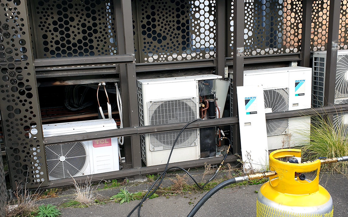 A grey recovery cylinder attached to three white air conditioning units on-site
