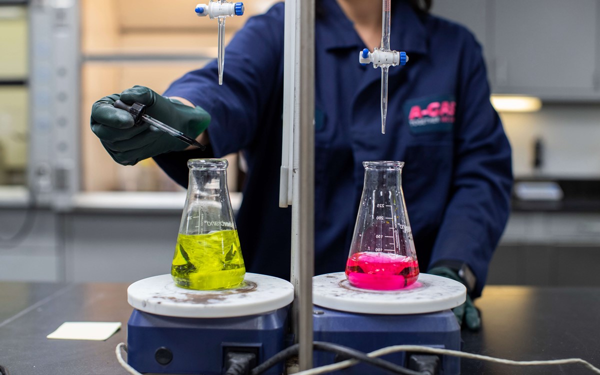 A woman carrying out laboratory analysis, she is wearing a navy lab coat and safety glasses, , one is yellow and one is pink