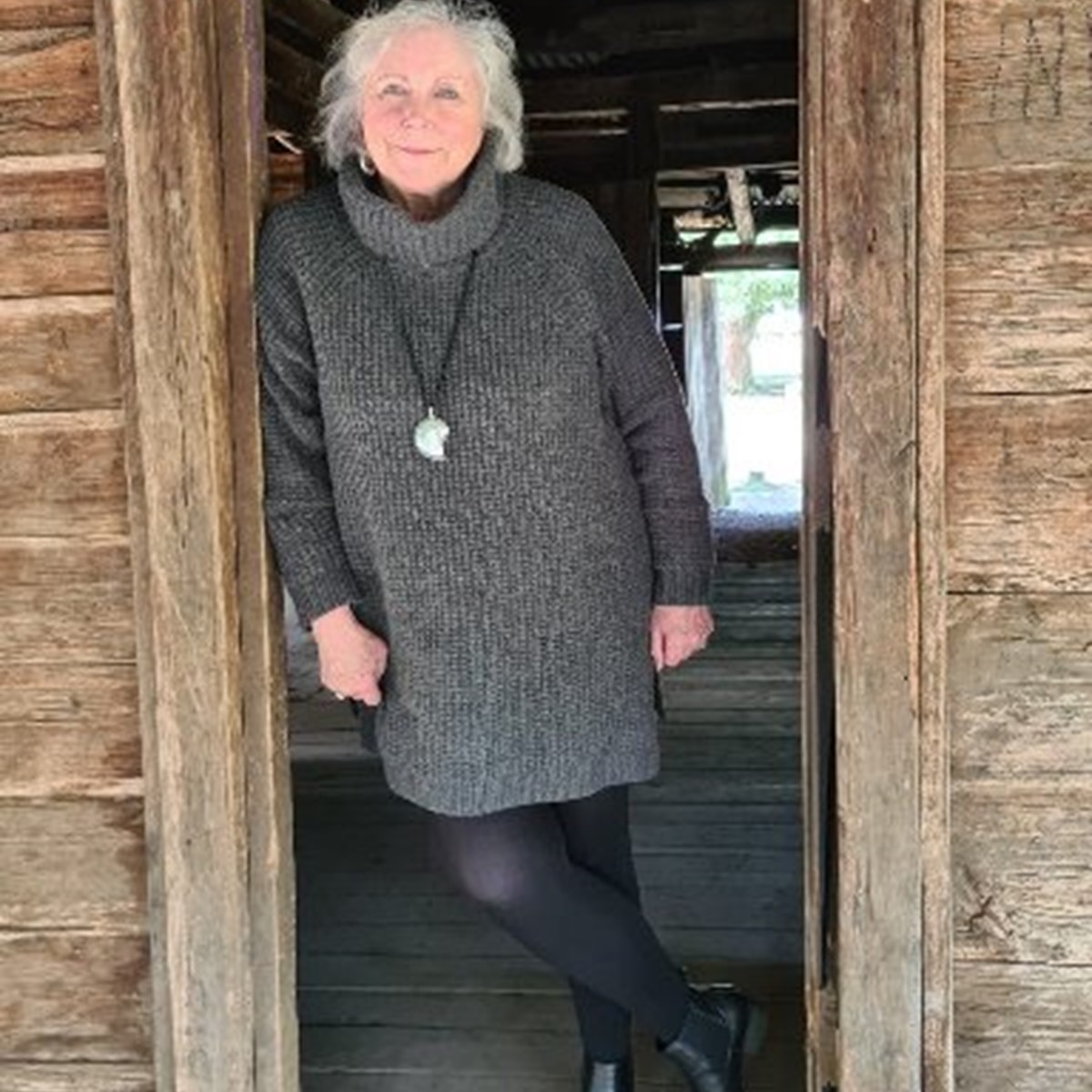 A woman in a grey dress standing in a doorway of a wooden building and smiling