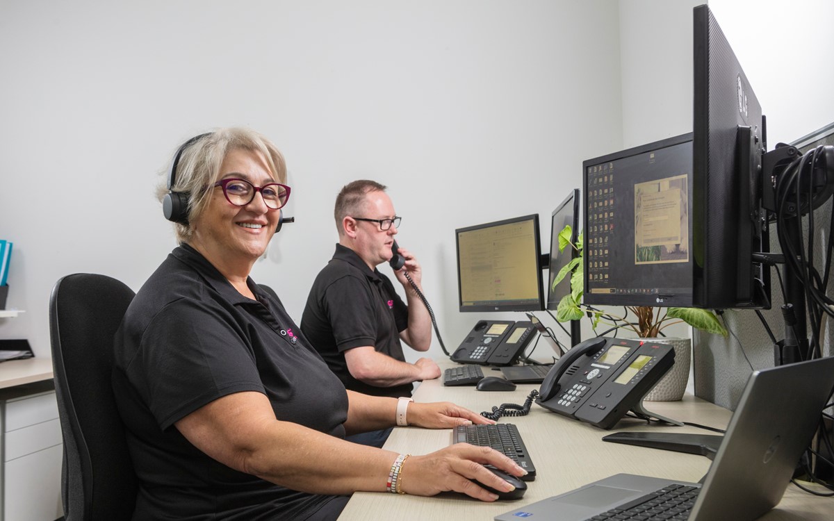 A woman wearing a headset smiling at the camera and a man on the telephone at a desk