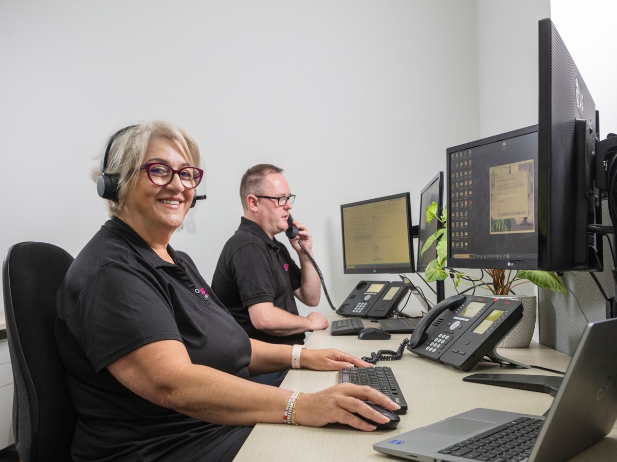 A woman wearing a headset smiling at the camera and a man on the telephone at a desk