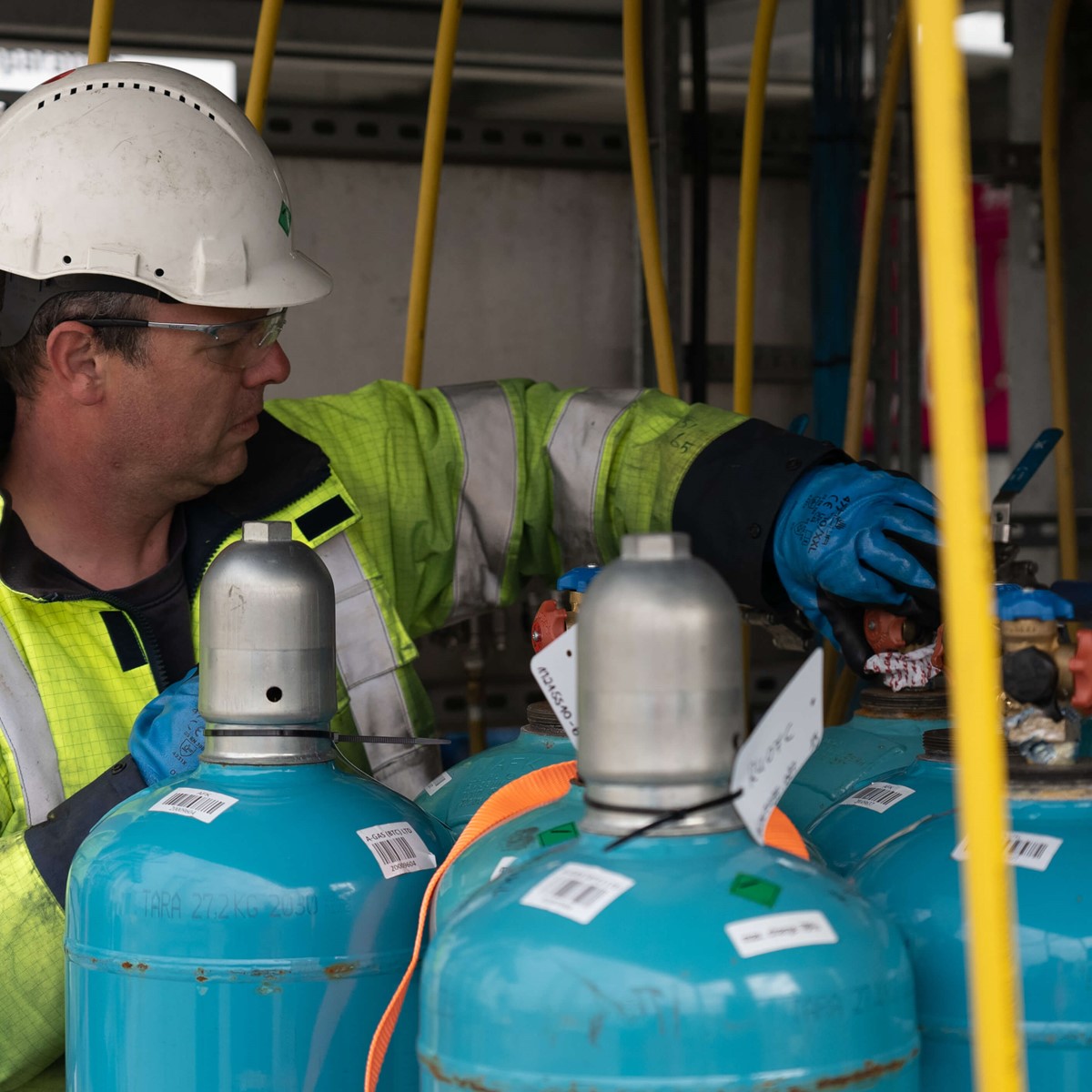 A man in a helmet and yellow hi-vis jacket works on blue cylinders