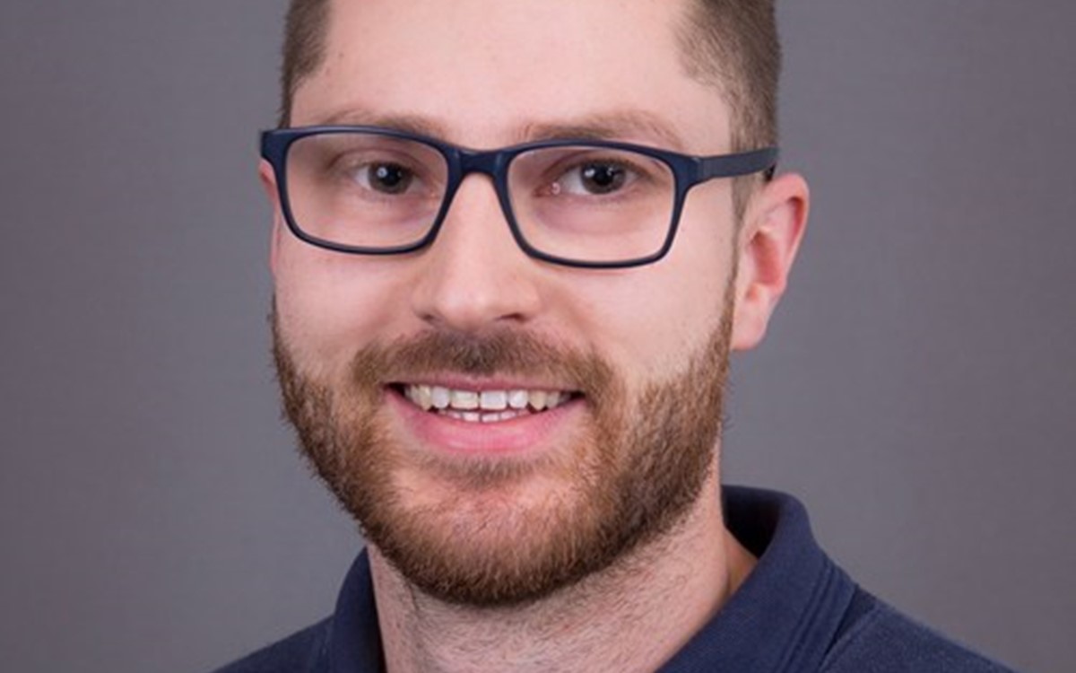 A headshot of a man in a blue polo and glasses smiling on a grey background