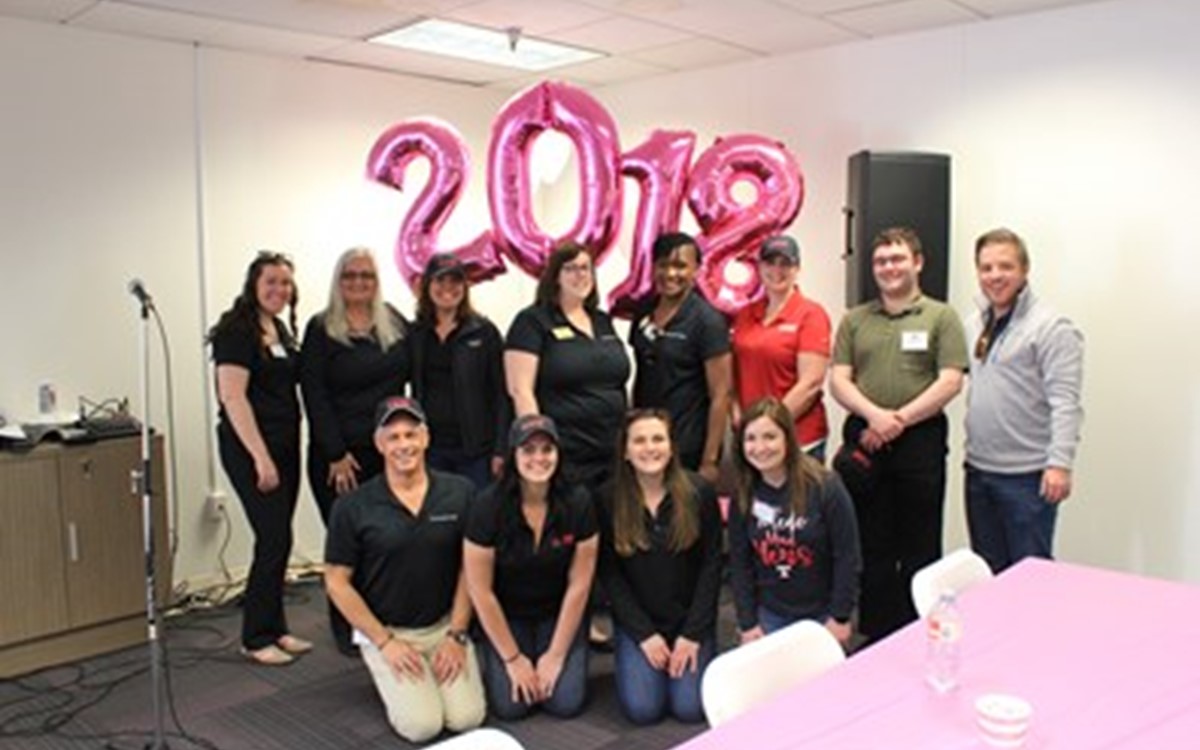 A group of people wearing dark blue polos in an office with pink balloons