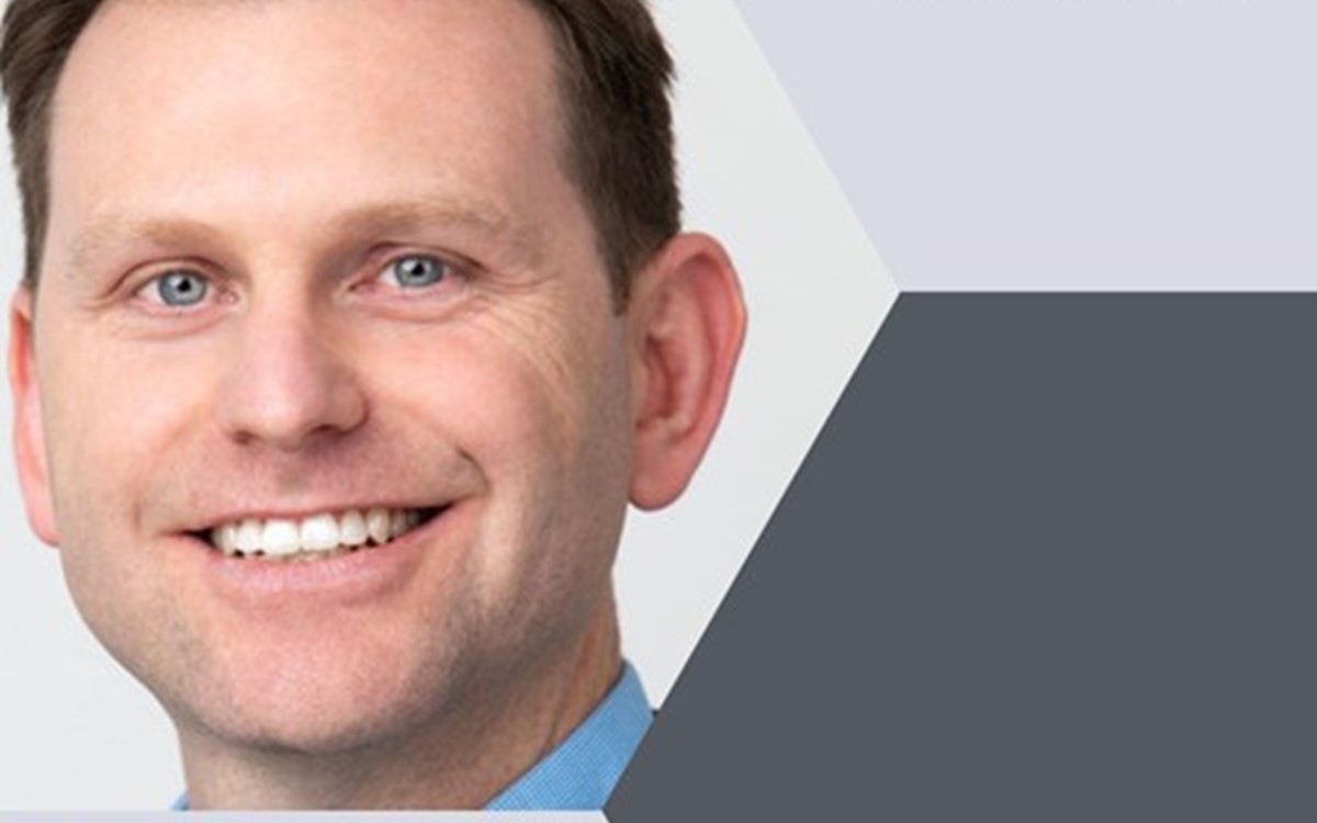 A man smiles at the camera on a background of light and dark grey hexagons