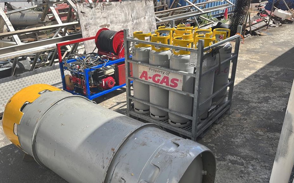 A large grey drum in front of a cage of grey cylinders with yellow tops on-site