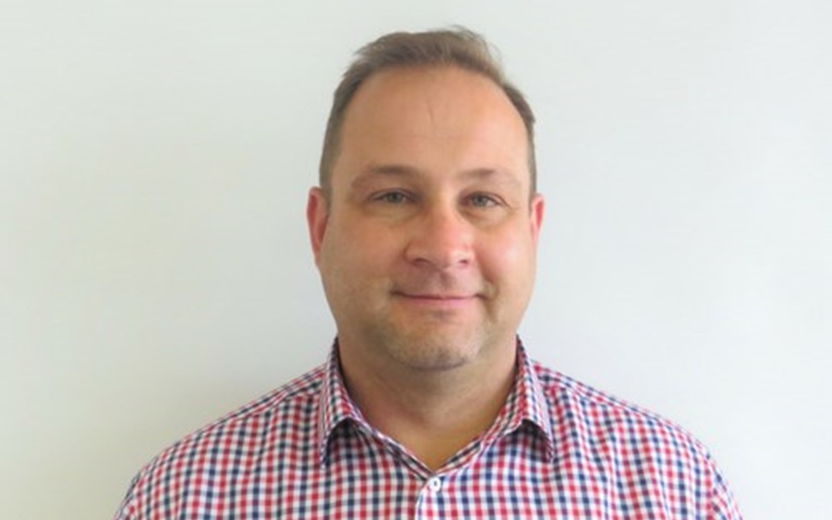 A man in a checked shirt smiles at the camera against a light grey background