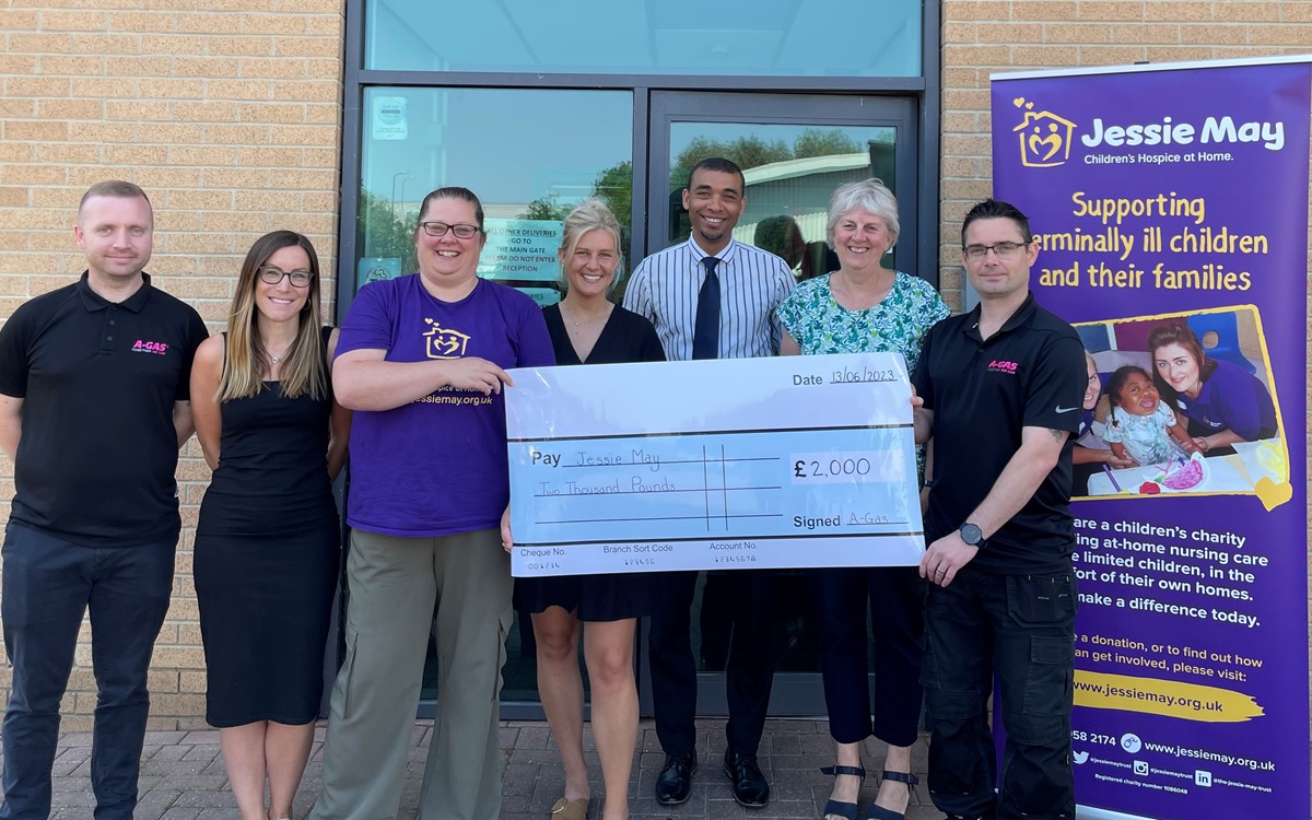 A group of people stand outside a building a hold a large white check next to a purple Jessie May banner