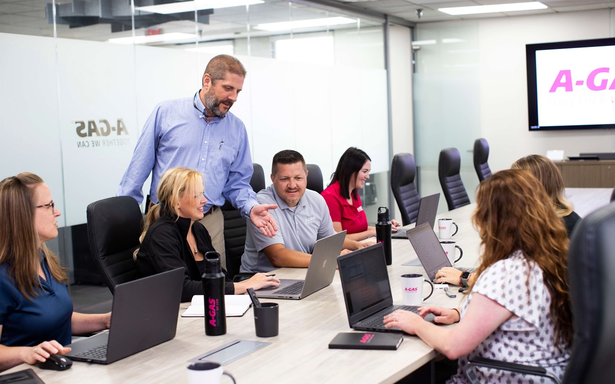 Men and women sat in the boardroom looking at a laptop, there is one man standing up pointing at the screen