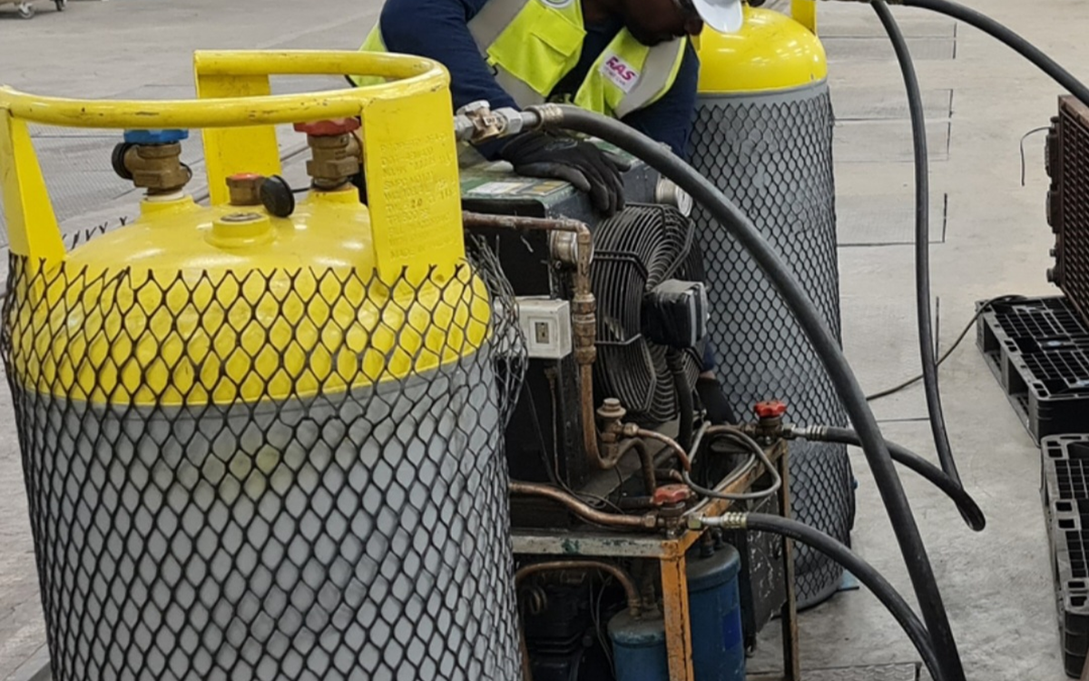 A man in a yellow high-vis jacket works with equipment between two large grey cylinders in a warehouse
