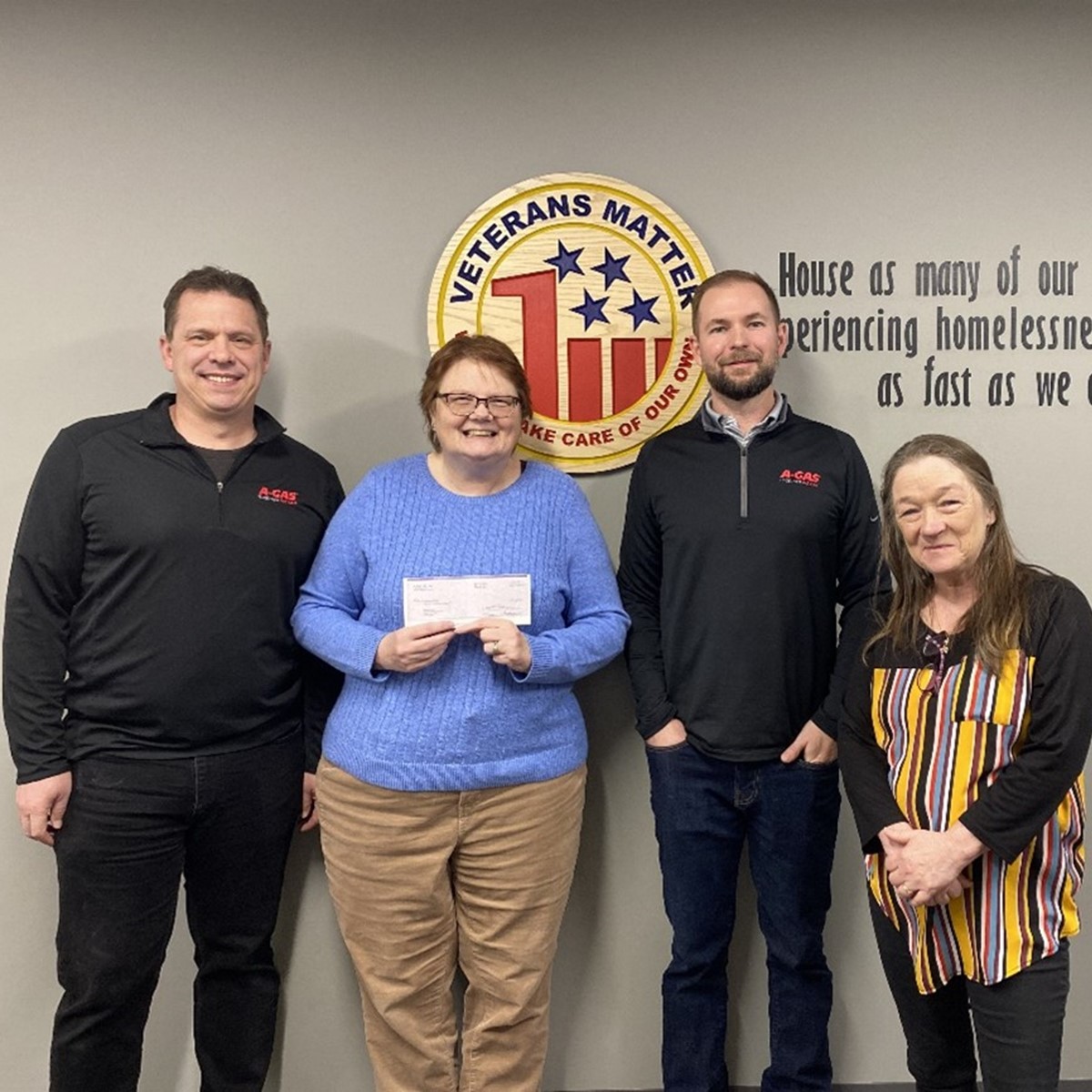 Four people stand holding a white check and smiling in front of a grey background and the "Veterans Matter" logo